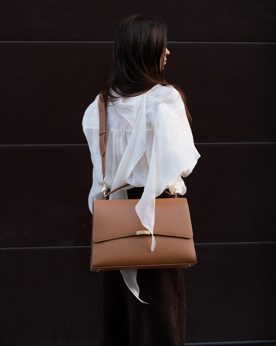 Person wearing a white blouse and holding a brown leather bag against a dark background