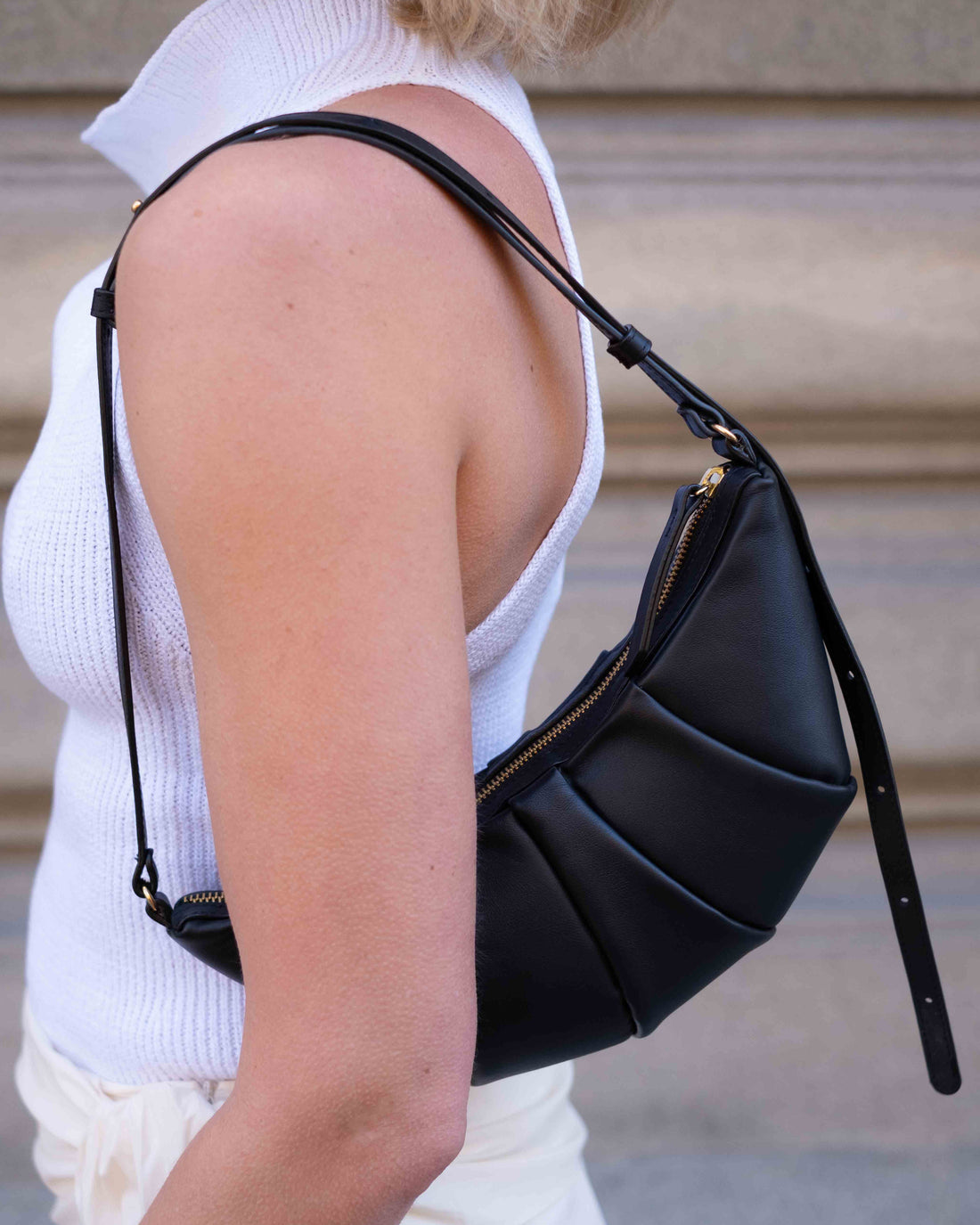 A black leather crescent-shaped shoulder bag with a long thin strap, featuring a gathered pleated front design, shown against a plain light gray background.