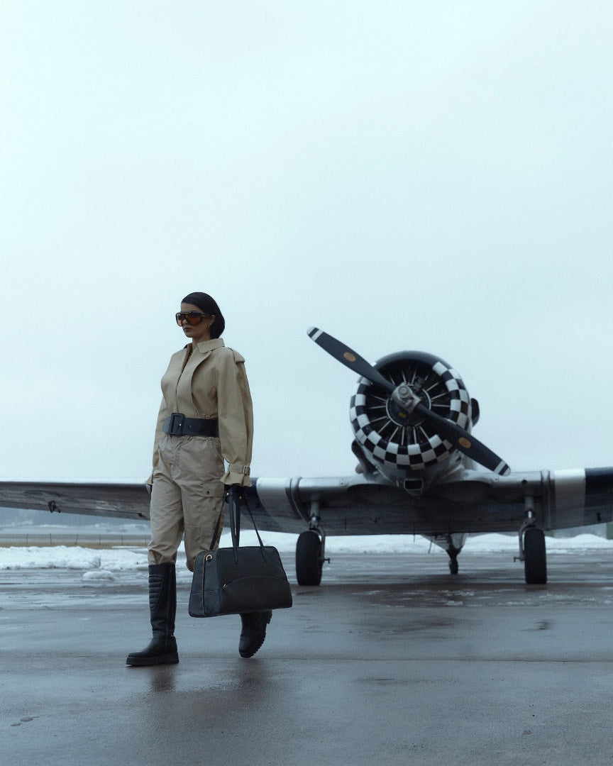 A woman walking from an airplane, carrying a black leather duffel bag with golden detailng.