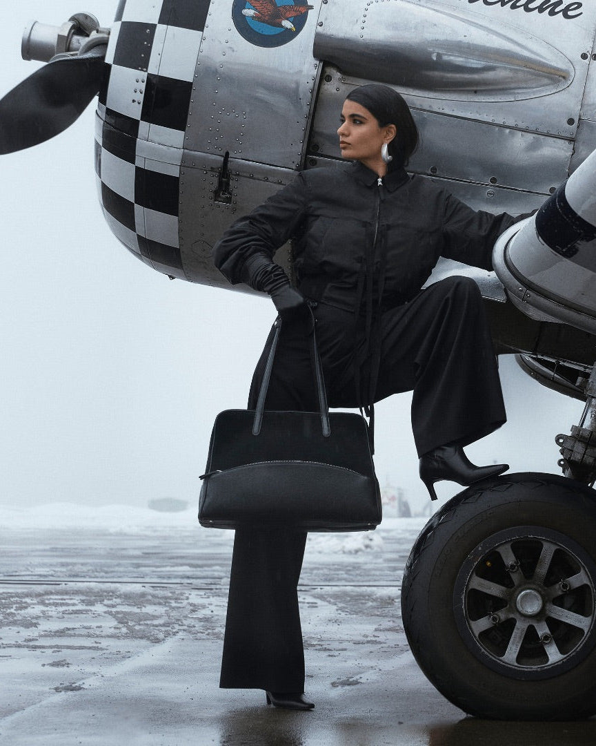 A woman on the outside of an airplane, holding a black suede and leather duffel bag with silver detailing. 
