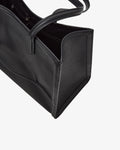 Close-up of a black pebbled leather tote bag with a top handle, showing the side panel and interior opening, and a luggage strap, against a light gray background.