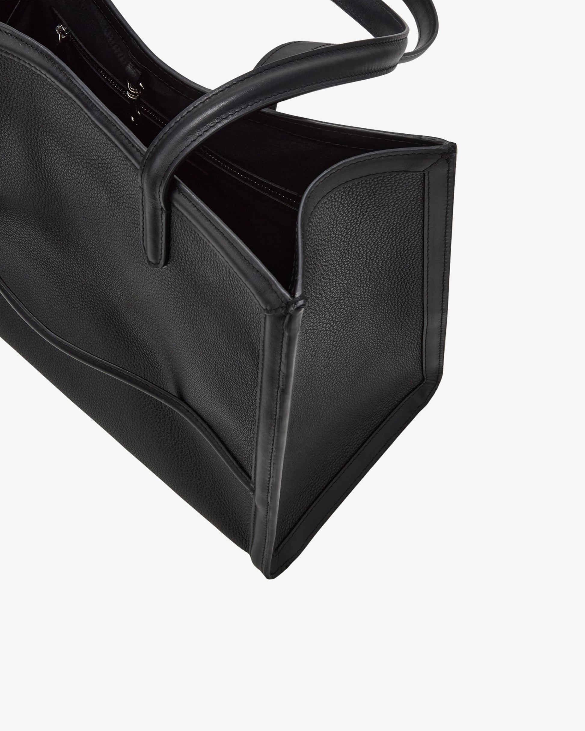 Close-up of a black pebbled leather tote bag with a top handle, showing the side panel and interior opening, and a luggage strap, against a light gray background.