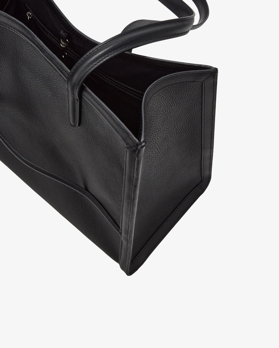 Close-up of a black pebbled leather tote bag with a top handle, showing the side panel and interior opening, and a luggage strap, against a light gray background.