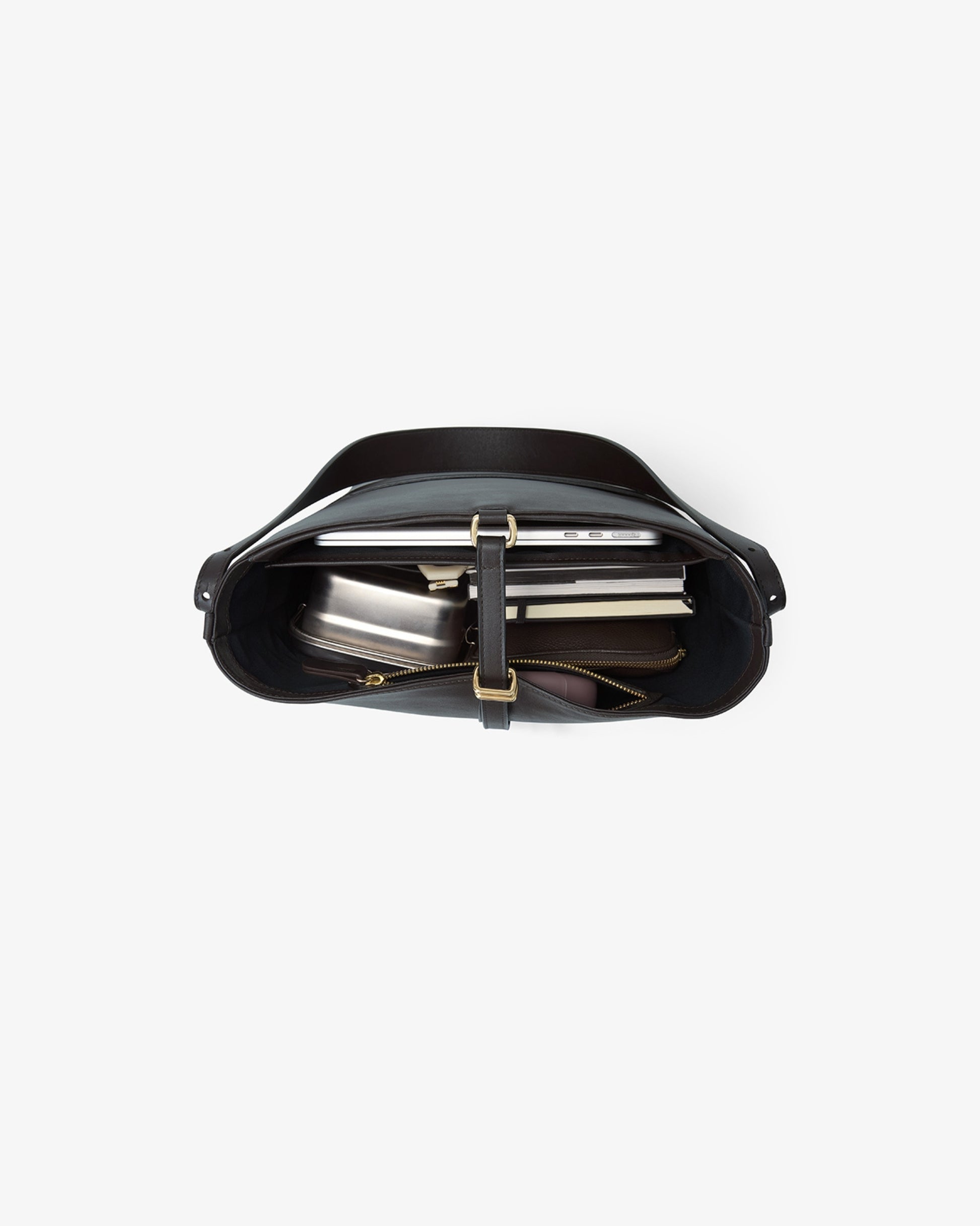 A top-down view of a black leather shoulder bag with a structured curved silhouette, featuring a central strap closure with a gold-tone buckle and a zippered pocket containing books or folders, set against a white background with thin red horizontal guideline markings.
